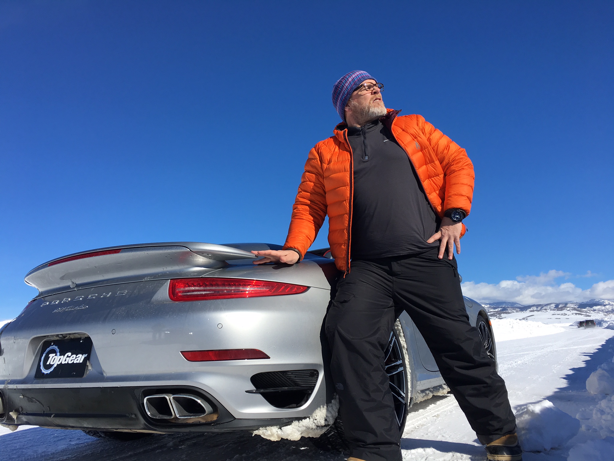 Donny in an orange jacket standing beside a silver Porsche on a snowy Top Gear USA location